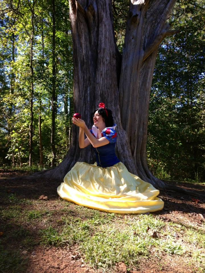 Performer in a blue and yellow fairytale gown holding a red apple beneath a large tree